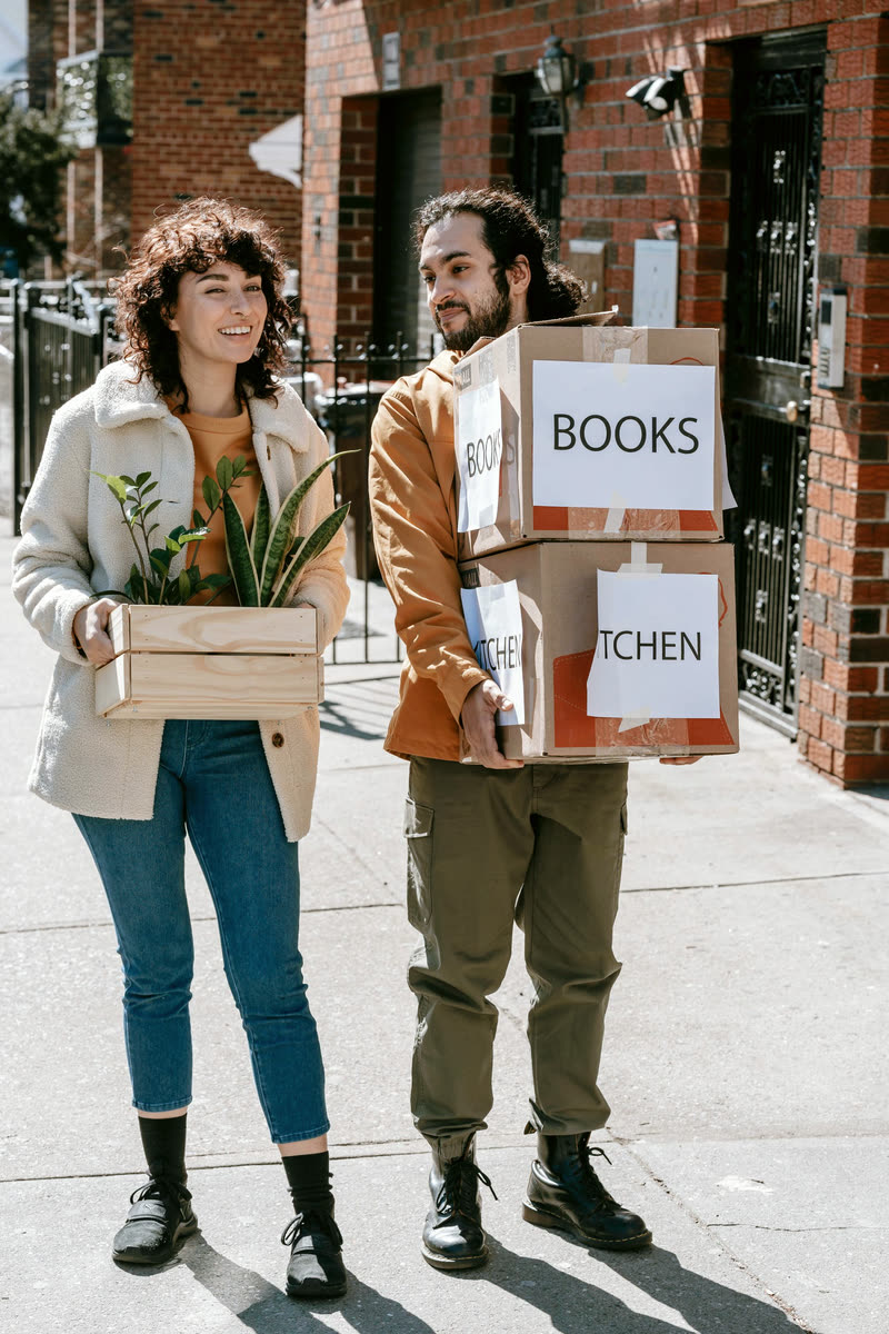 Two people arriving together carrying moving boxes and a plant